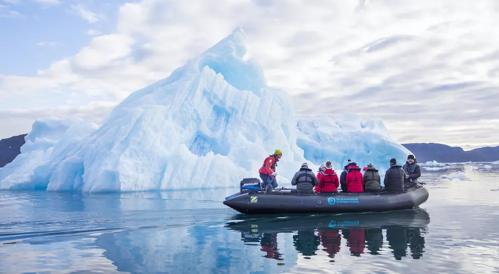 A group of people in a boat near a large blue iceberg on calm water under a cloudy sky.
