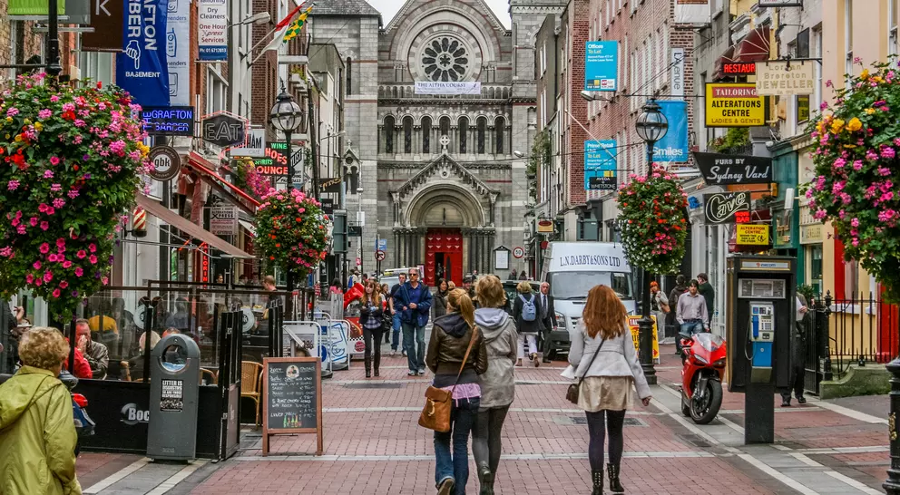 Bustling city street lined with shops and colorful flowers, leading to a historic church in the background.