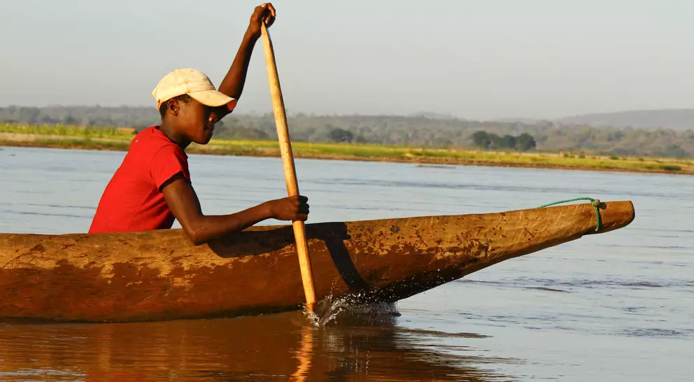 A person in a red shirt paddles a wooden canoe on a calm river during daylight.