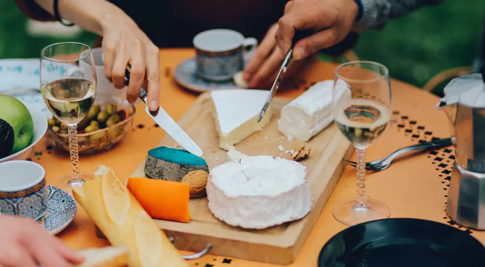 A close-up of a cheese platter with various cheeses, wine glasses, and fruits on a table. Hands are cutting cheese.