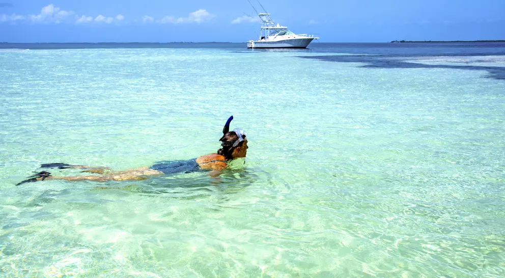 A person snorkels in clear turquoise water, with a boat in the background and a blue sky above.
