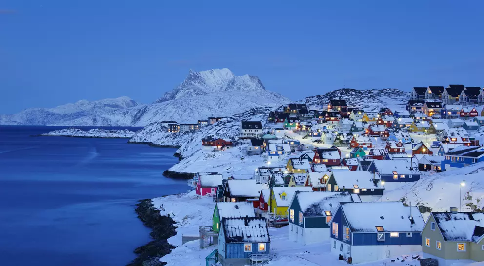 Colorful snowy houses line a coastal hill, with mountains in the background and a serene blue twilight sky.