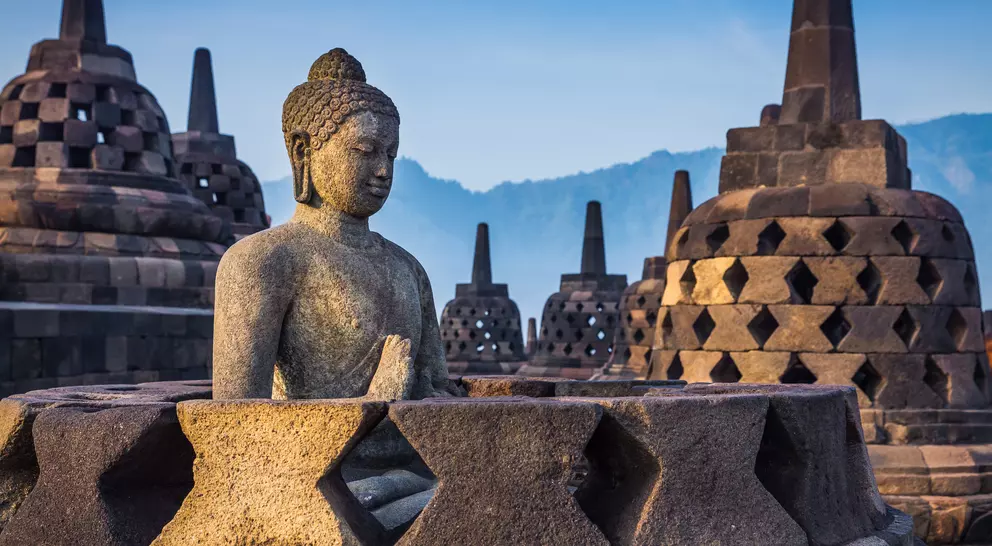 Buddha statue in the foreground with ancient, tiered stupas against a hazy mountain backdrop at sunrise.