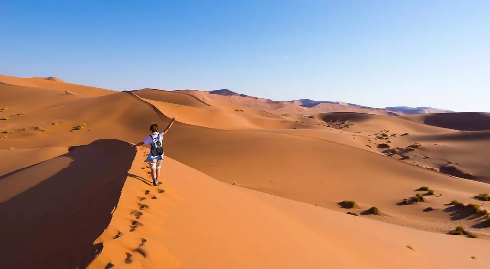 A person walks on a sandy dune in a desert, with rolling hills and a clear blue sky in the background.