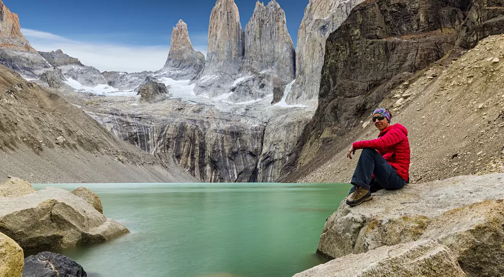 Person in red sitting on rocks by a turquoise lake, with towering mountains in the background under a clear blue sky.