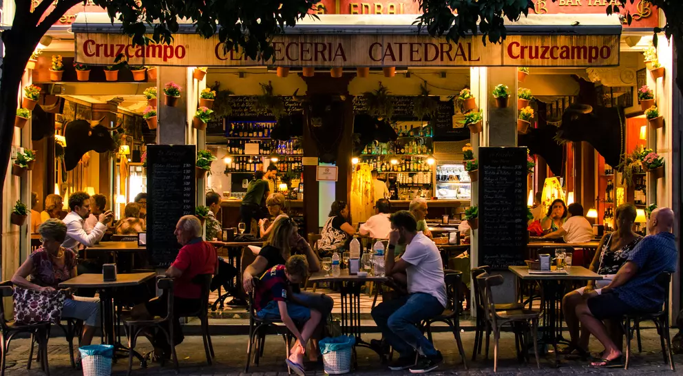 A lively outdoor café with people dining at tables, illuminated by warm lights, surrounded by greenery and a welcoming atmosphere.
