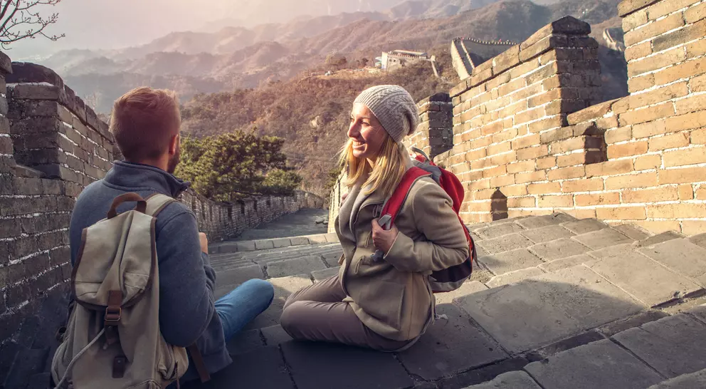 Two hikers sit on the Great Wall of China, enjoying the view of mountains in the background during sunset.