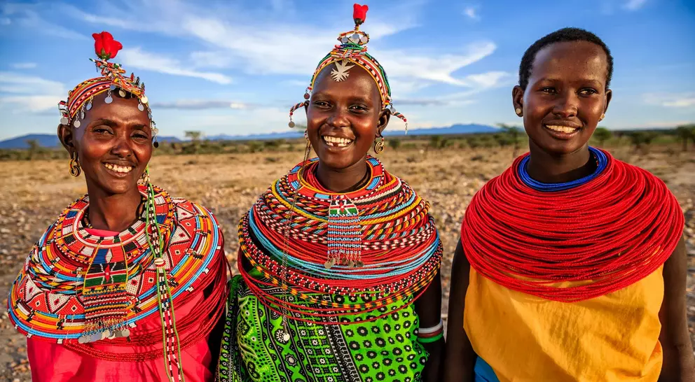 Three smiling women in colorful traditional attire stand together against a scenic landscape.
