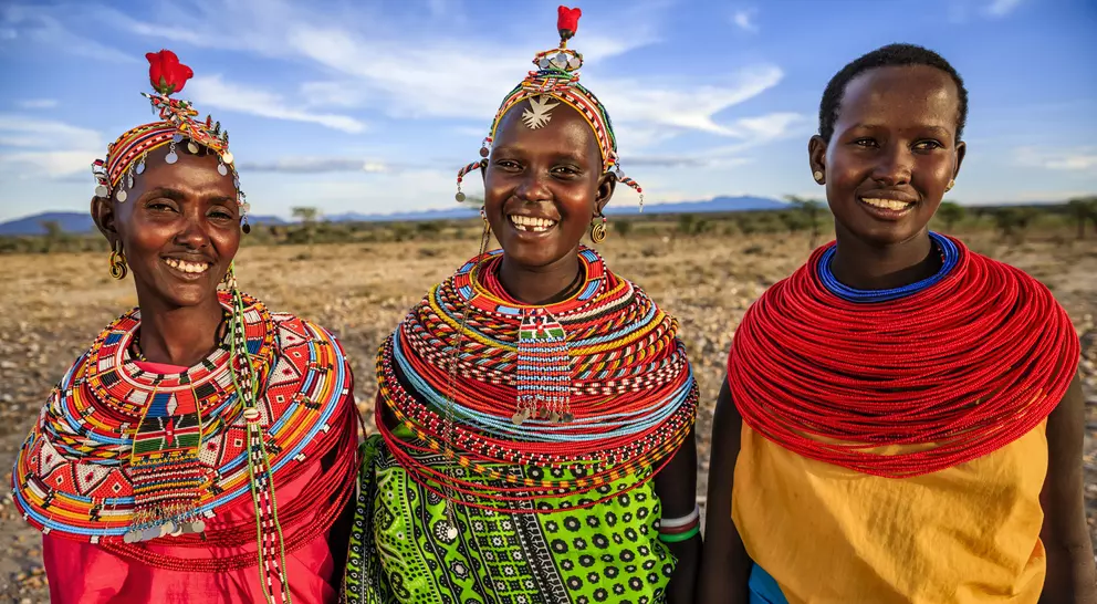 Three smiling women in colorful traditional attire stand together against a scenic landscape.