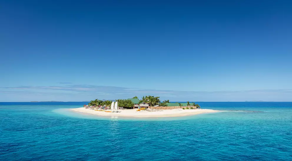Beautiful small island in the middle of the south pacific ocean with beach huts, lounge chairs, palm trees, surrounded with beautiful clear turquoise water.