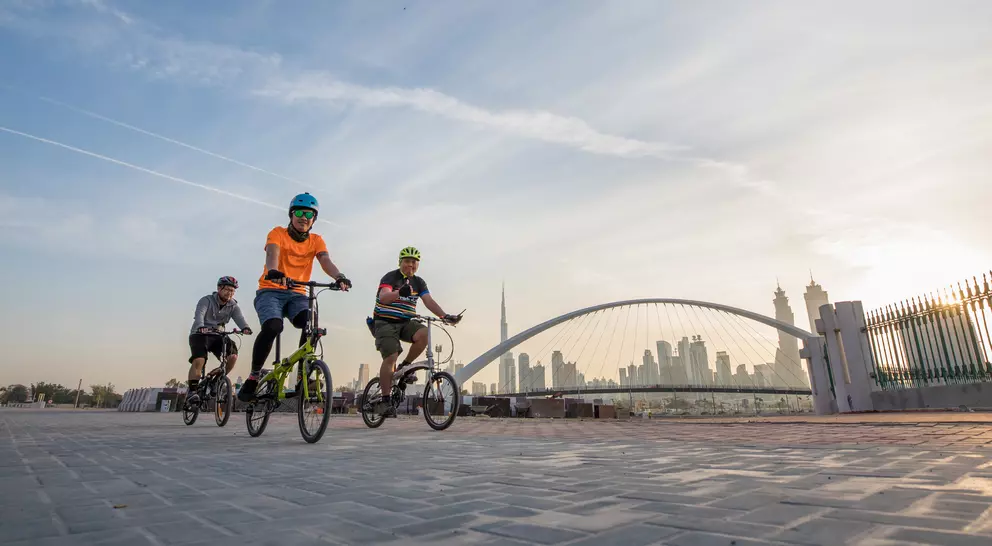 Three children ride bicycles along a path with a city skyline and bridge in the background under a sunset sky.