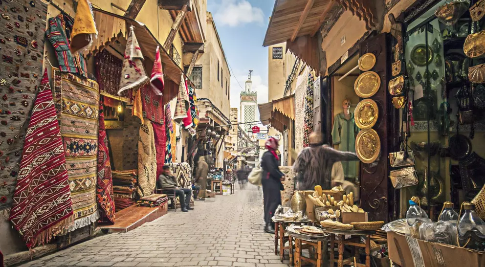 Narrow market street lined with colorful rugs, shops, and people browsing various goods under a clear blue sky.