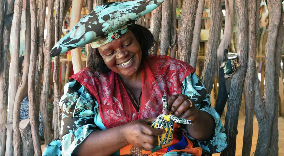 Smiling woman in colorful attire crafts a decorative doll, surrounded by natural wooden elements in a vibrant setting.