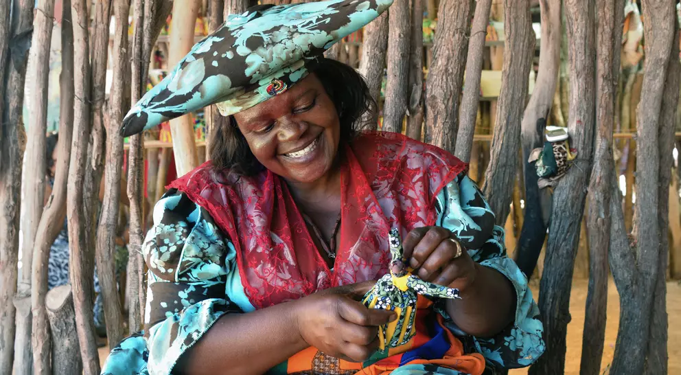 Smiling woman in colorful attire crafts a decorative doll, surrounded by natural wooden elements in a vibrant setting.