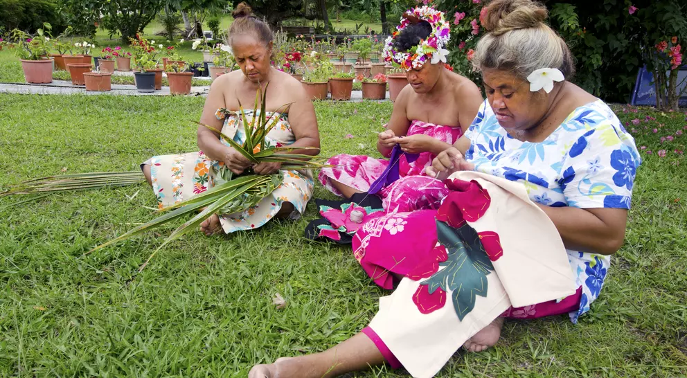 Three women sitting on grass, weaving with leaves and fabric, surrounded by colorful flowers and plants in pots.