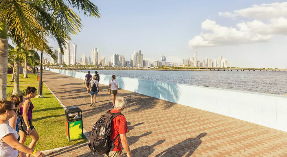 People walking along a waterfront promenade with a skyline and palm trees in the background under a clear sky.