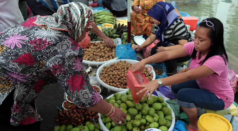 A vibrant market scene with women selling fruits from baskets along a waterway, showcasing various colorful produce.
