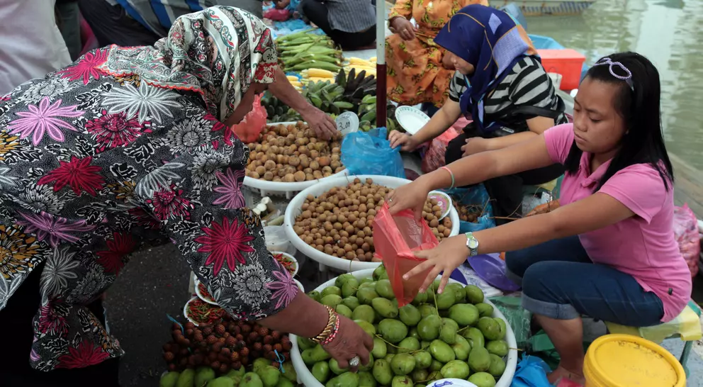 A vibrant market scene with women selling fruits from baskets along a waterway, showcasing various colorful produce.