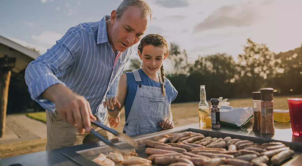 An adult man and a girl are grilling sausages together at a barbecue, enjoying the outdoor setting.