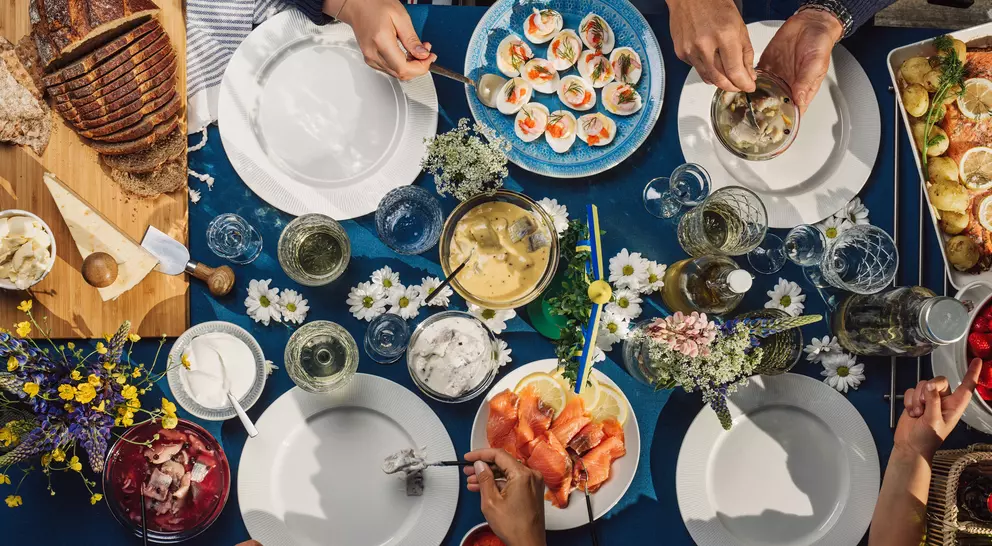 Aerial view of a vibrant outdoor table set with various dishes, drinks, and floral decorations, with hands reaching for food.