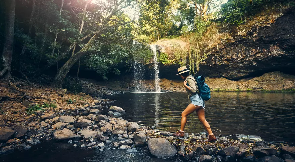 A person in shorts and a hat walks along rocks by a forest stream, with a waterfall in the background.