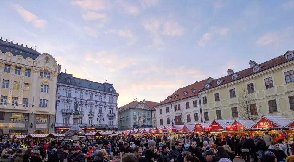 Crowded market scene with stalls under a colorful sky, surrounded by historic buildings.