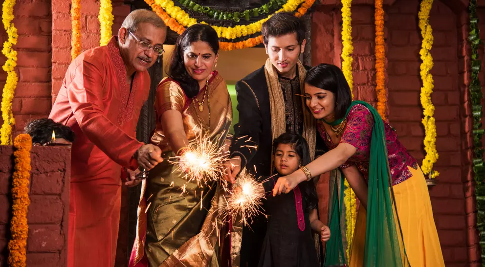 A family celebrates together, lighting sparklers in traditional attire surrounded by colorful flowers and festive decorations.