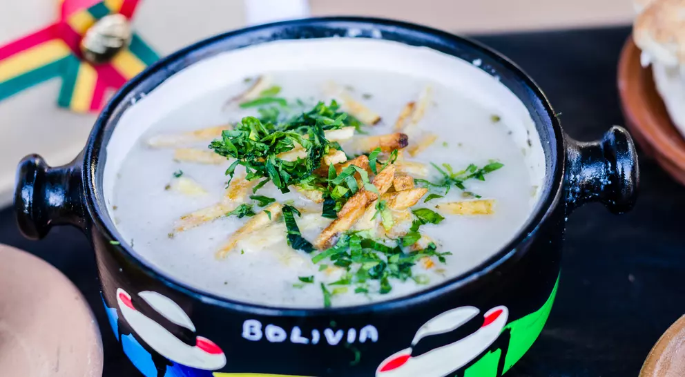 A colorful bowl of traditional Bolivian soup topped with herbs and crispy strips, alongside bread on a wooden surface.
