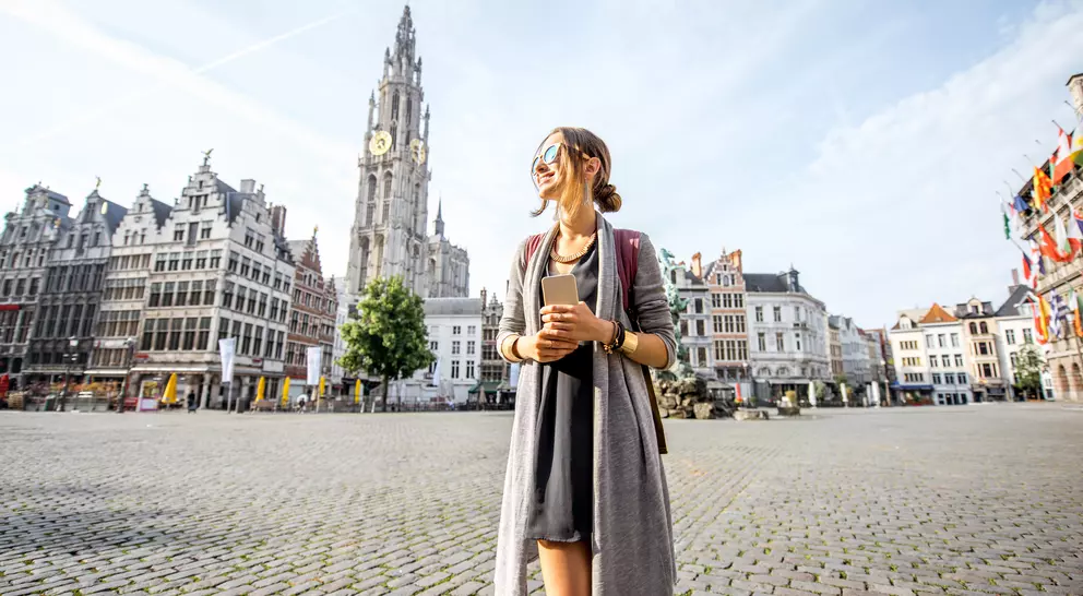 A woman stands in a plaza with historic buildings and a clock tower in the background, enjoying a sunny day.