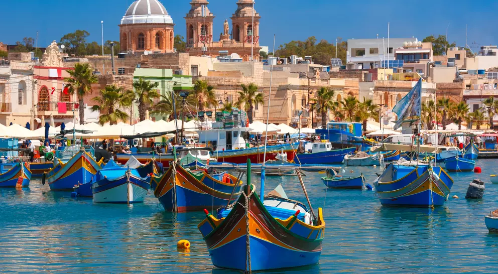 Colorful boats in a vibrant harbor, with historic buildings and palm trees under a bright blue sky.
