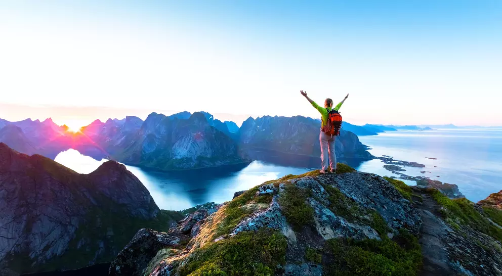 A person with arms raised stands on a rocky peak, overlooking a colorful landscape of mountains and water at sunrise.