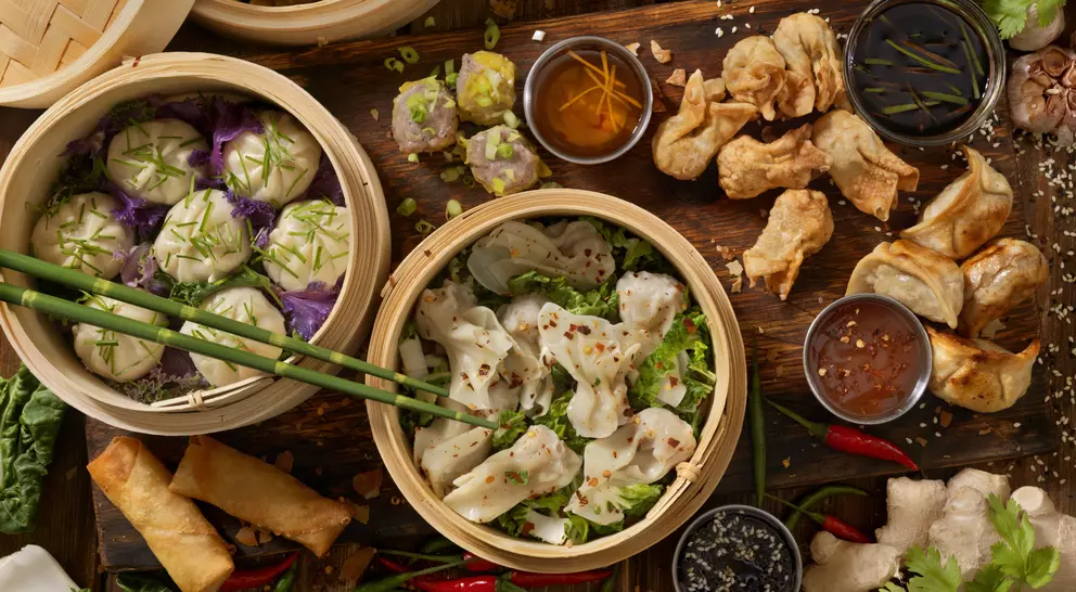 A spread of various Asian dumplings and spring rolls, served in bamboo steamers and accompanied by dipping sauces.