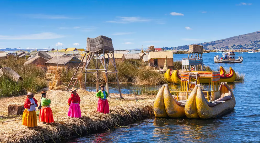 Colorful traditional boats and local women in vibrant clothing by the floating islands on a lake under a clear blue sky.