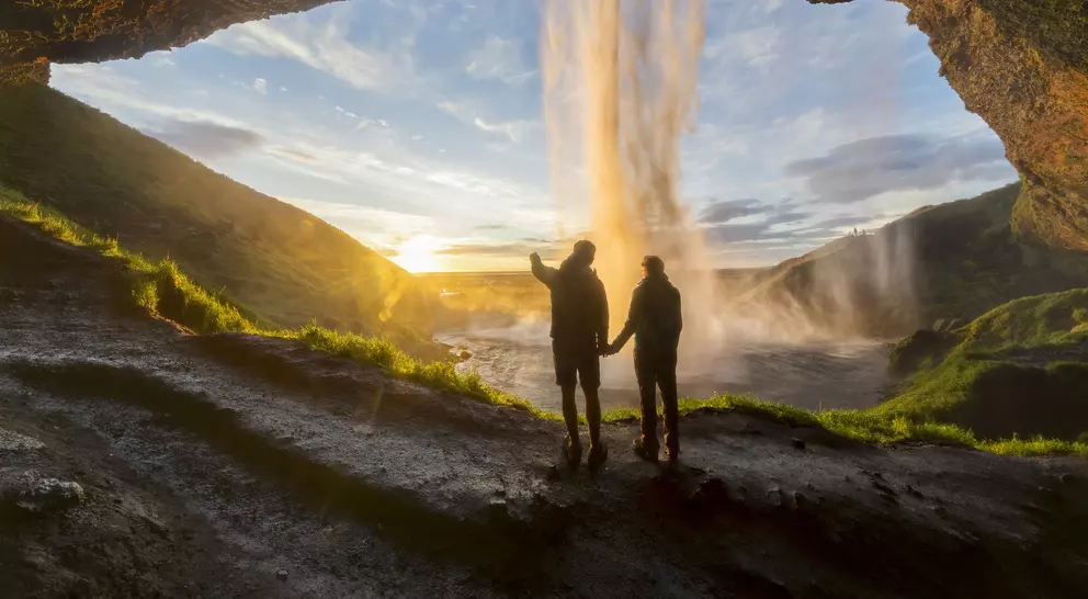 Silhouetted figures stand hand in hand under a waterfall, framed by a rocky cavern and a colorful sunset.