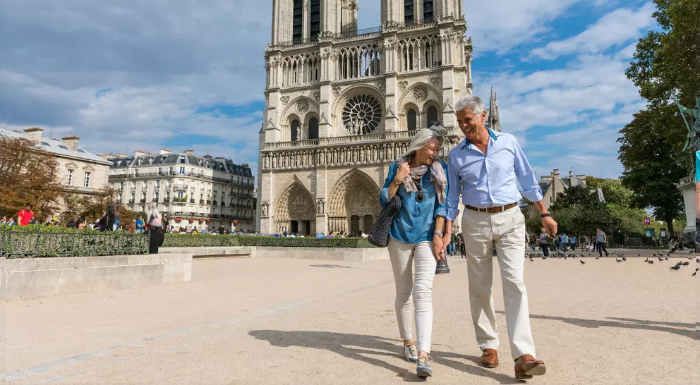 A couple walks hand-in-hand in front of Notre-Dame Cathedral on a sunny day, with people and trees in the background.