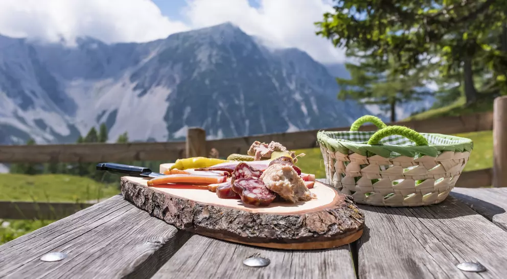 A rustic wooden table holds a charcuterie platter with cured meats and vegetables, set against a scenic mountain backdrop.