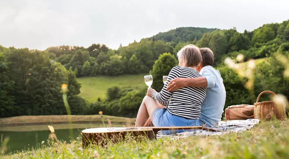 A couple sits together by a serene lake, enjoying a drink and the view of green hills and trees in the background.