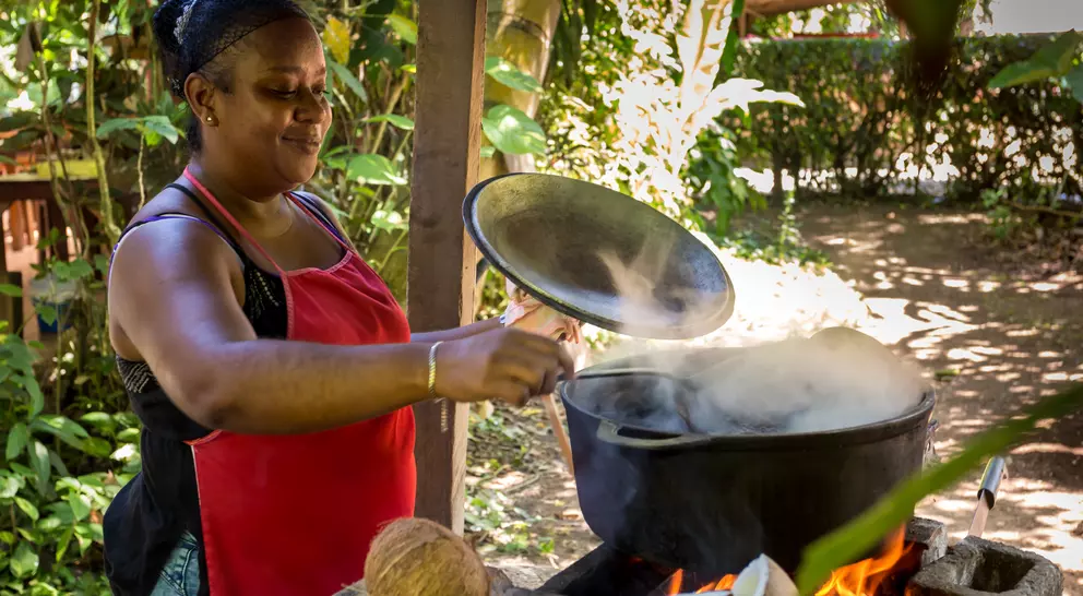 A woman in a red apron stirs a pot over a fire outdoors, surrounded by greenery and coconuts.