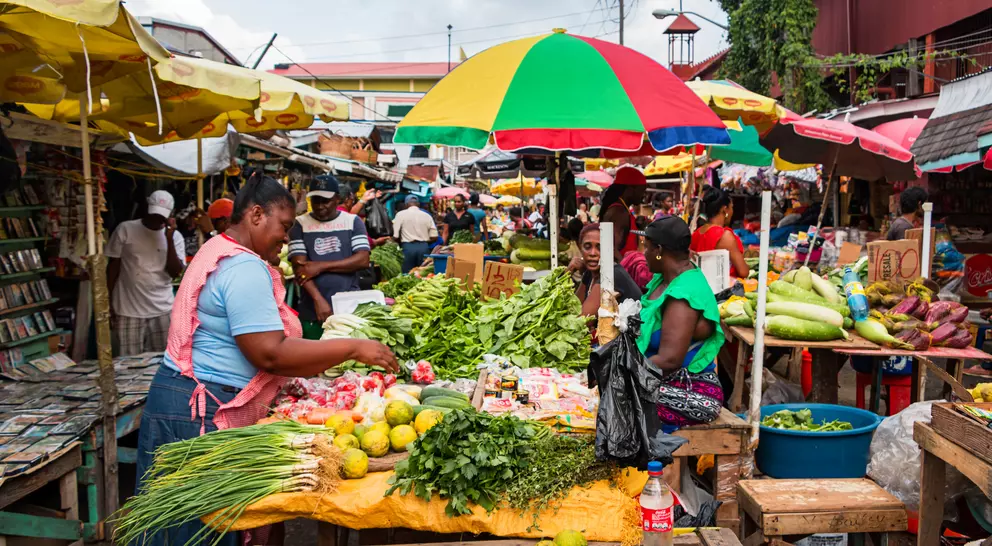 A vibrant market scene featuring vendors selling fresh produce under colorful umbrellas, with shoppers browsing the stalls.