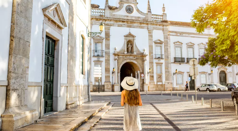 A woman in a hat walks towards a historic building, surrounded by trees and sunlight in a charming street setting.