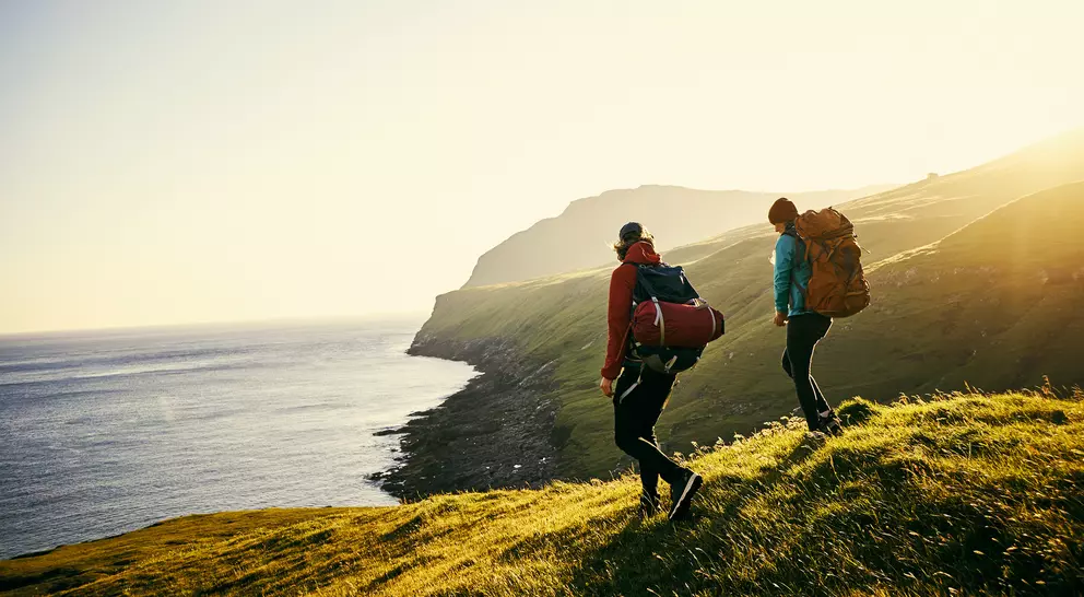 Two hikers with backpacks walk along a grassy hillside, overlooking a calm sea under a bright sunset.