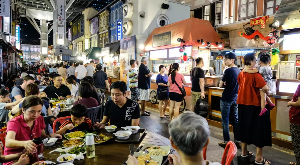 Busy night market scene with people dining and standing near food stalls, surrounded by colorful shopfronts.