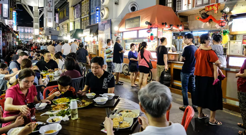 Busy night market scene with people dining and standing near food stalls, surrounded by colorful shopfronts.