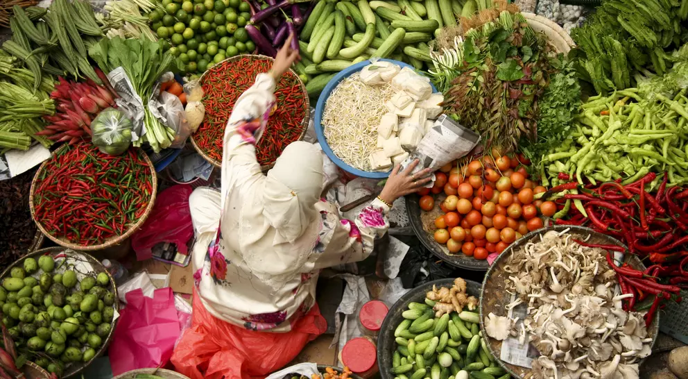 A vendor in a market reaches for produce among vibrant displays of fruits and vegetables in various baskets.