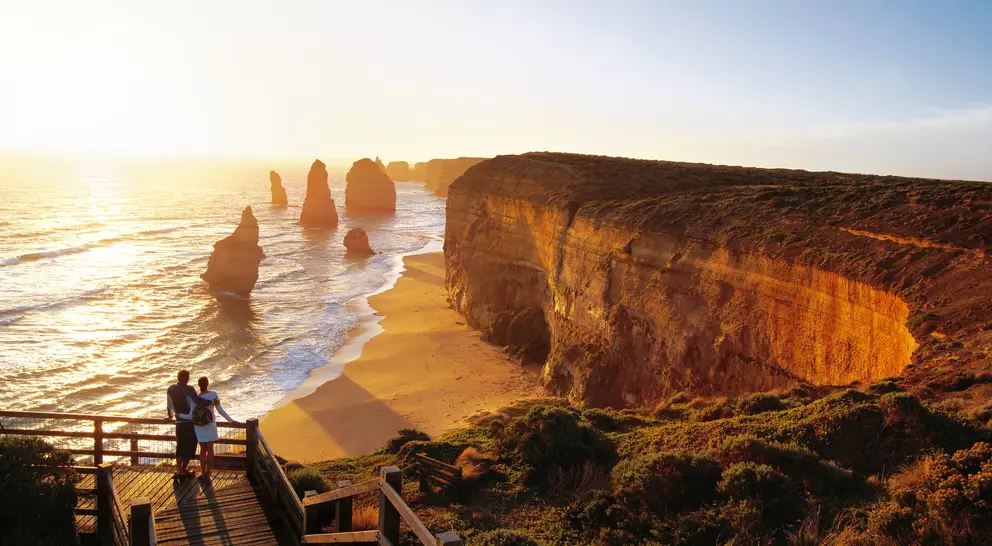A couple stands on a wooden walkway overlooking a beach with cliffs and sea stacks at sunset.