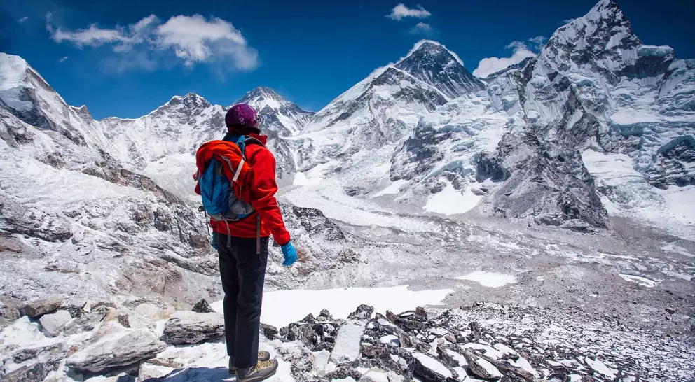 Person in a red jacket stands on rocky terrain, overlooking snow-capped mountains under a clear blue sky.