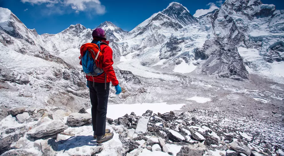 Person in a red jacket stands on rocky terrain, overlooking snow-capped mountains under a clear blue sky.