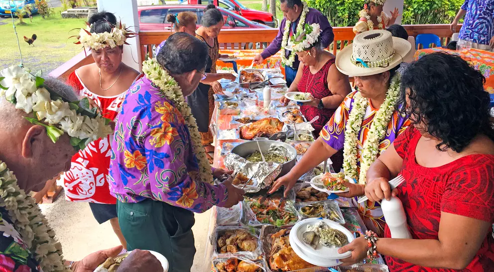 A group of people in colorful attire share a buffet spread outdoors, adorned with leis, enjoying a festive meal together.