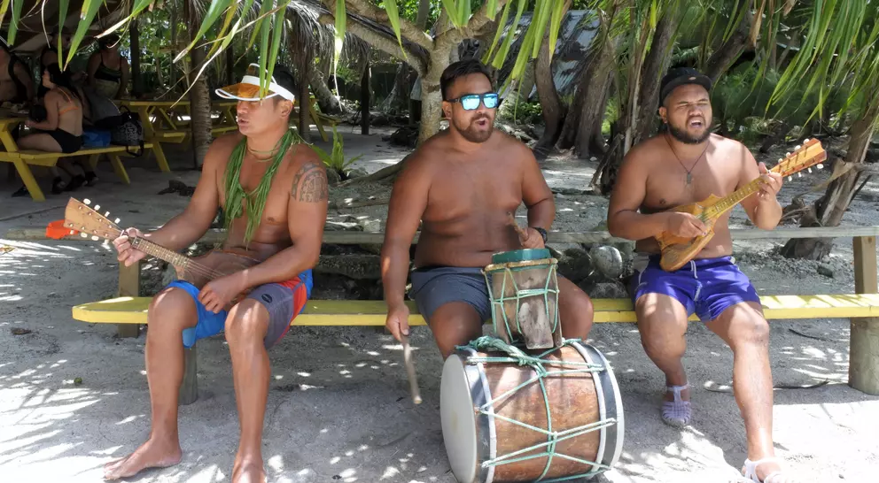 Three shirtless men play traditional instruments, with palm trees and a sandy beach in the background.