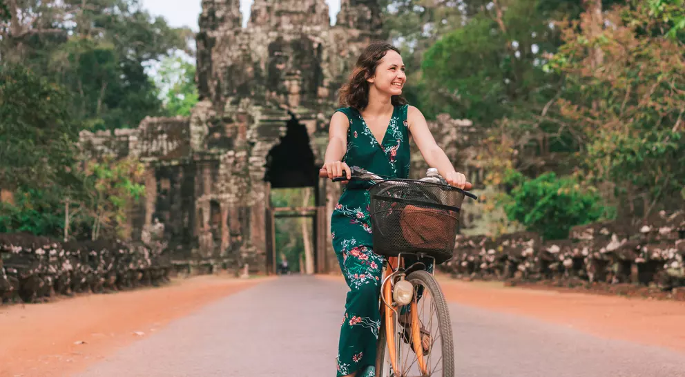 A woman rides a bicycle on a path near a historic stone temple surrounded by lush greenery.